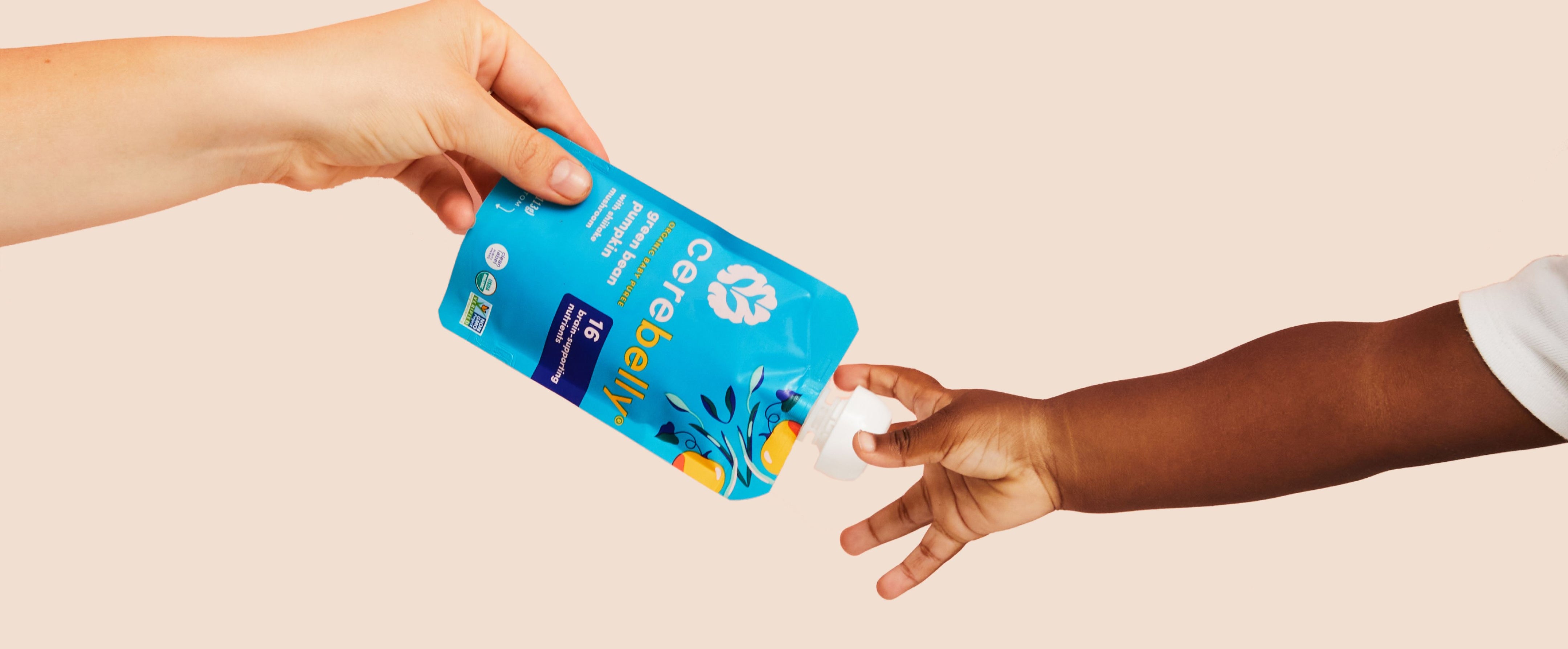 Hand passing a green bean pumpkin pouch to another hand on a beige background